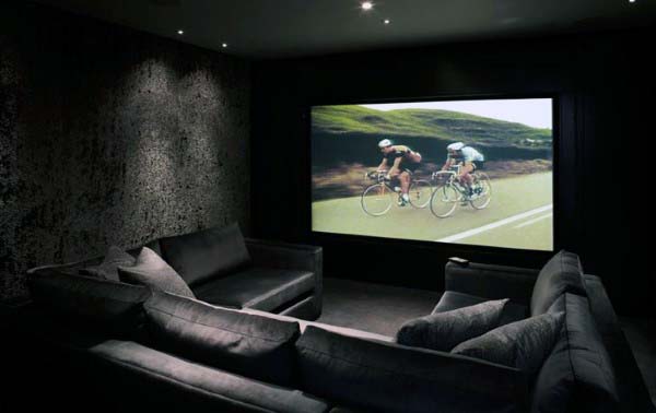 Home theater with a large screen showing cyclists on a road, surrounded by plush, dark-hued sofas