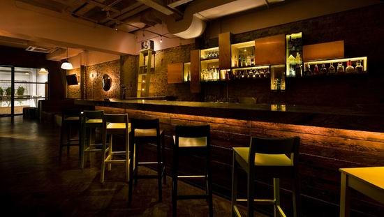 Dimly lit bar with empty stools against a sleek counter, shelves of bottles glowing behind