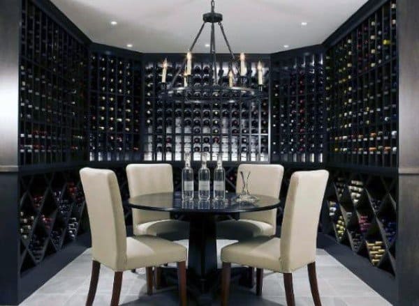 A chandelier hangs over a table and four chairs in a wine cellar, with bottles lining the black shelves
