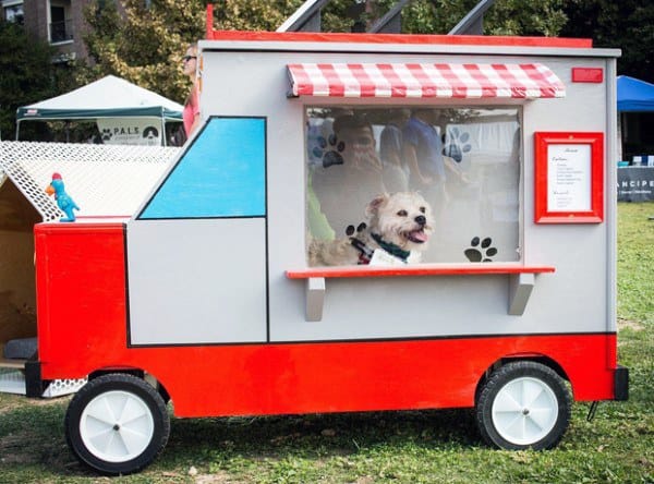 Dog sitting inside a food truck-themed dog house with a striped awning and a window featuring paw prints, set in an outdoor area