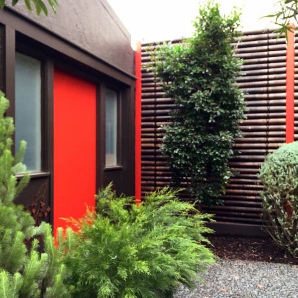 Metal-framed horizontal bamboo fence in a modern courtyard with vibrant plants and a red-accented building.