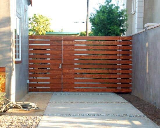 Wooden slat gate between concrete walls with a small driveway, trees in the background