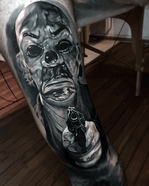 Realistic black and grey tattoo of a sinister masked figure pointing a gun, featuring intense shading and eerie details for a chilling horror effect.