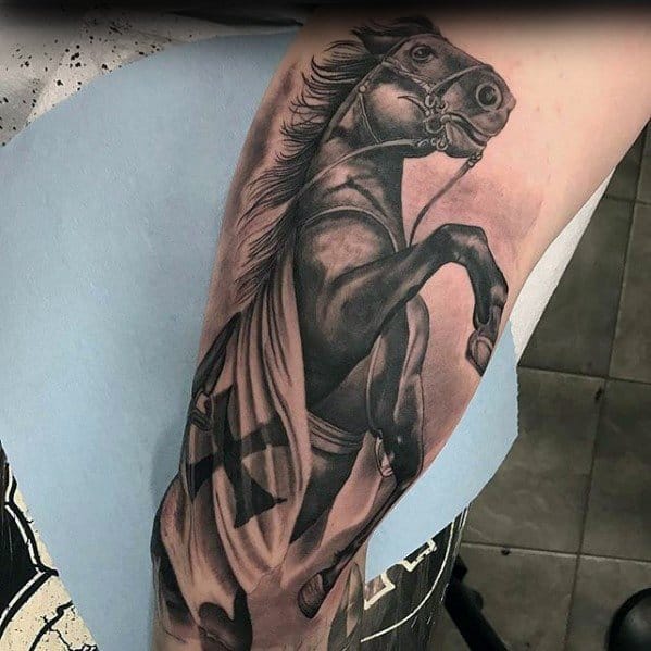 Black and grey tattoo of a powerful rearing horse draped in a crusader-style cloth, placed on the upper arm with detailed shading