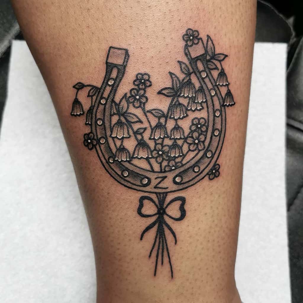 Black ink tattoo of a horseshoe adorned with Lily of the Valley flowers, small blossoms, and a bow