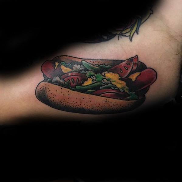 Hot dog tattoo featuring sausage, lettuce, tomatoes, and mustard in a bun, with bold and vibrant details on the upper arm.