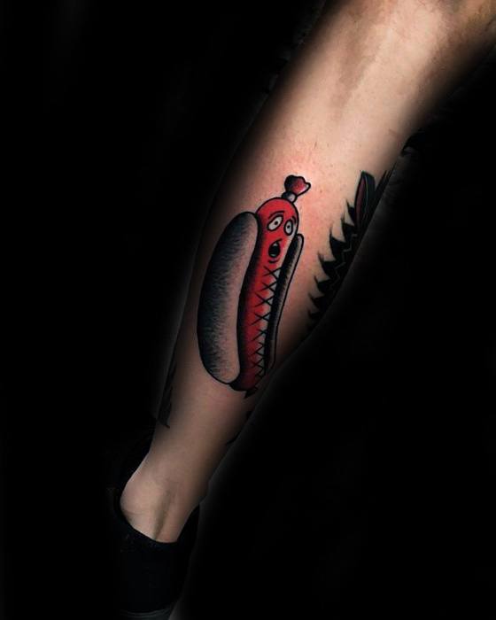 Whimsical hot dog tattoo featuring a red sausage with a smiling face, wrapped in a bun, on the calf.