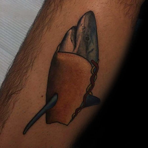 Unique tattoo of a fish inside a hot dog bun, with mustard drizzled over it, on the forearm.