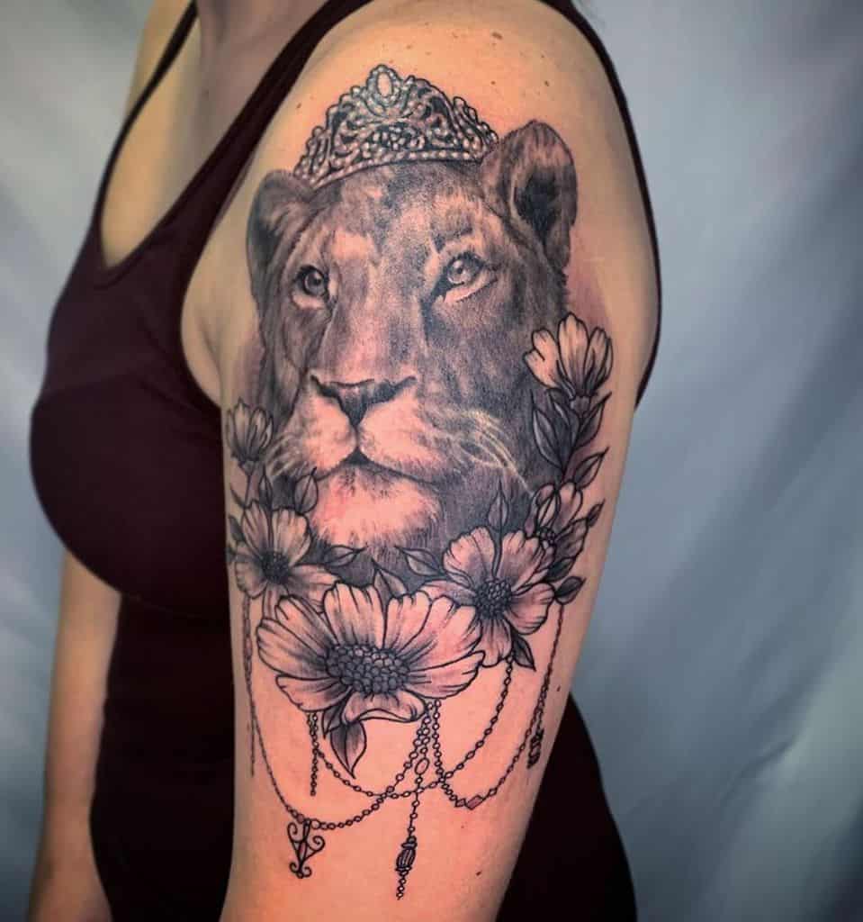 Black and grey lioness tattoo on the upper arm, featuring a regal tiara, floral details, and ornamental chains for a majestic and elegant look.