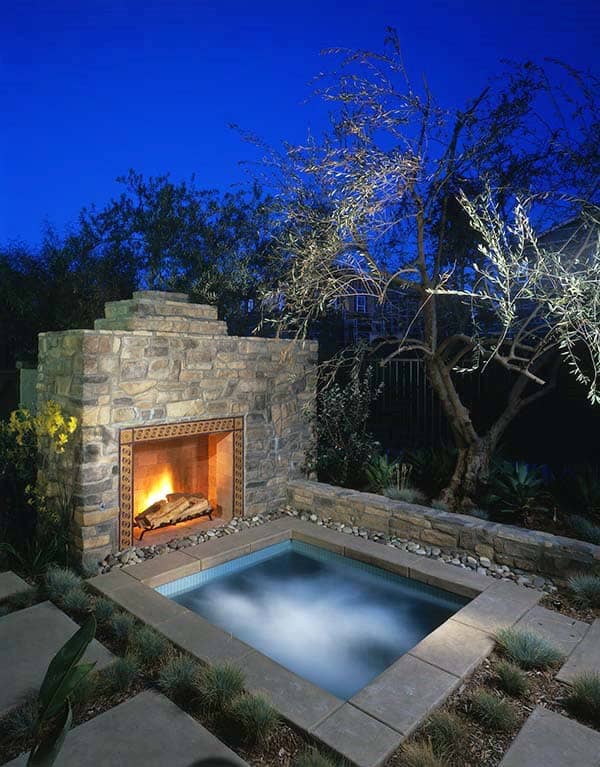Outdoor fireplace and lit pool surrounded by plants at dusk