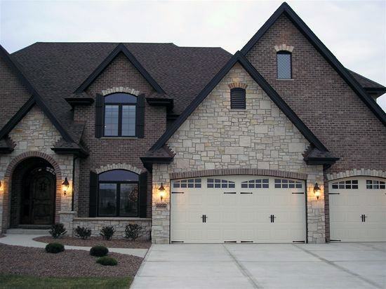 Modern home with dark brick and stone cladding, gabled rooflines, arched windows, and a three-car garage with illuminated sconces
