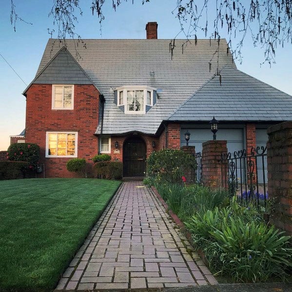 Brick walkway leading to a charming house with a slate roof, surrounded by lush green grass and neatly trimmed hedges