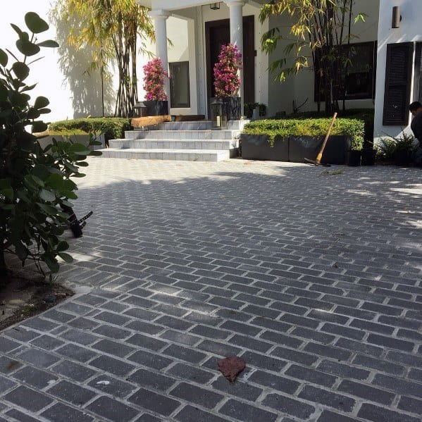 Brick-patterned driveway leading to a modern home entrance with greenery and decorative planters.