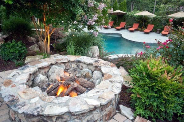 Stone fire pit near a pool with lounge chairs and landscaped greenery