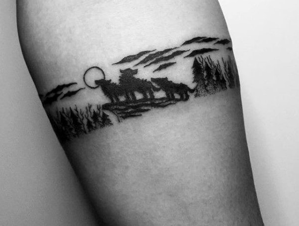 A black tattoo depicts a forest scene with silhouettes of wolves howling at the moon