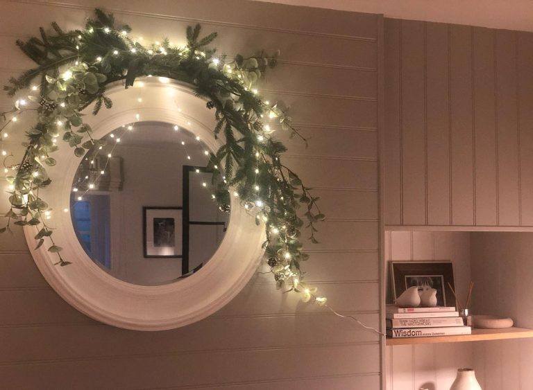 Round white-framed mirror decorated with fairy lights and greenery, reflecting a cozy, softly lit room with warm holiday charm.