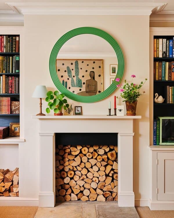Green-framed round mirror above a fireplace with stacked wood, plants, bookshelves, and colorful decor for a cozy yet vibrant look.