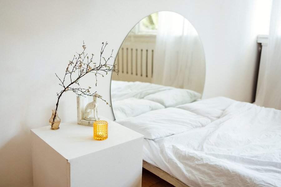 Arched mirror behind a white bed, reflecting soft natural light, with a minimalist nightstand featuring a branch in a vase and a warm candle.