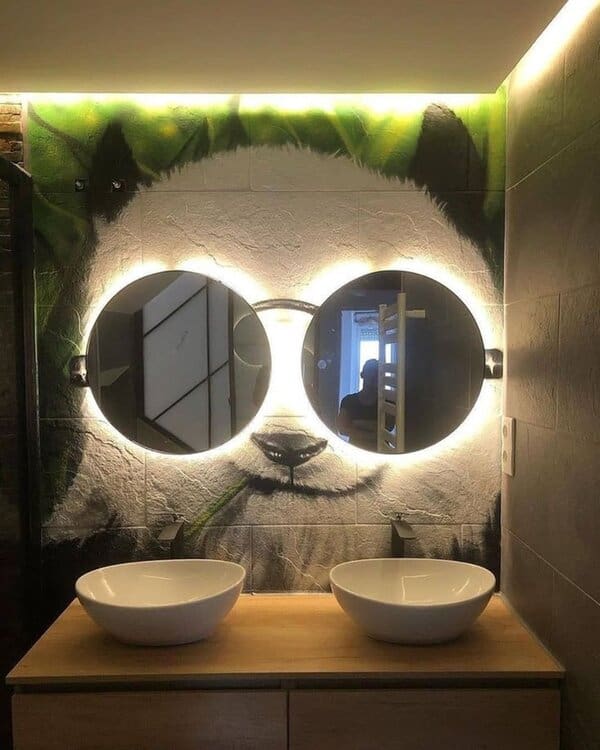 Backlit round mirrors over a panda mural in a modern bathroom with vessel sinks and wooden cabinetry for a playful yet stylish design.