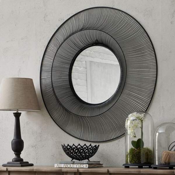 Black wire-frame round mirror on a textured wall, styled with a lamp, books, and glass-cloche orchids for a modern yet organic look.