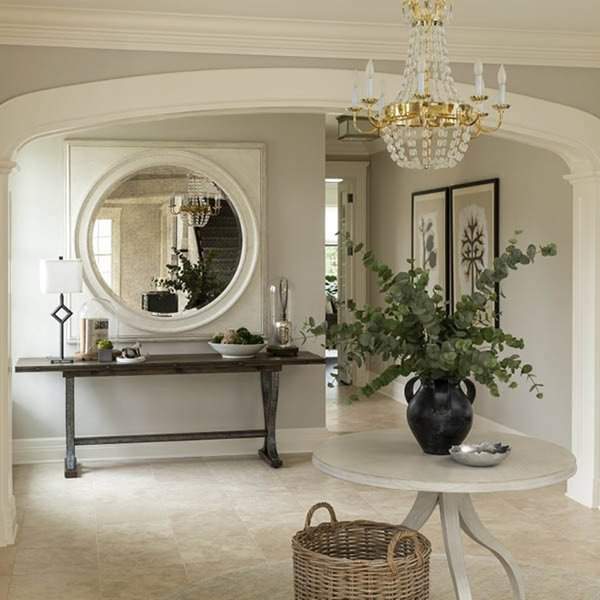 Large round mirror in a grand entryway, reflecting a crystal chandelier, with a rustic console table, greenery, and elegant neutral decor.
