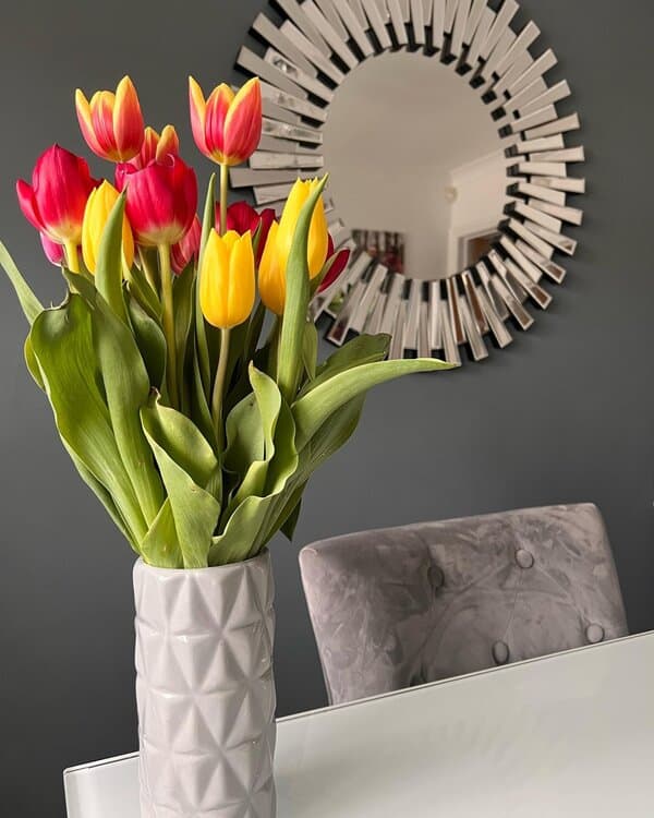 Sunburst mirror above a table with a bouquet of red and yellow tulips in a vase, set against a dark wall.