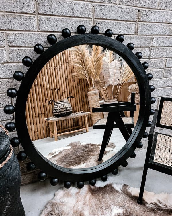 Large black-framed mirror with rounded details reflecting a boho-style space with bamboo, woven decor, and pampas grass.