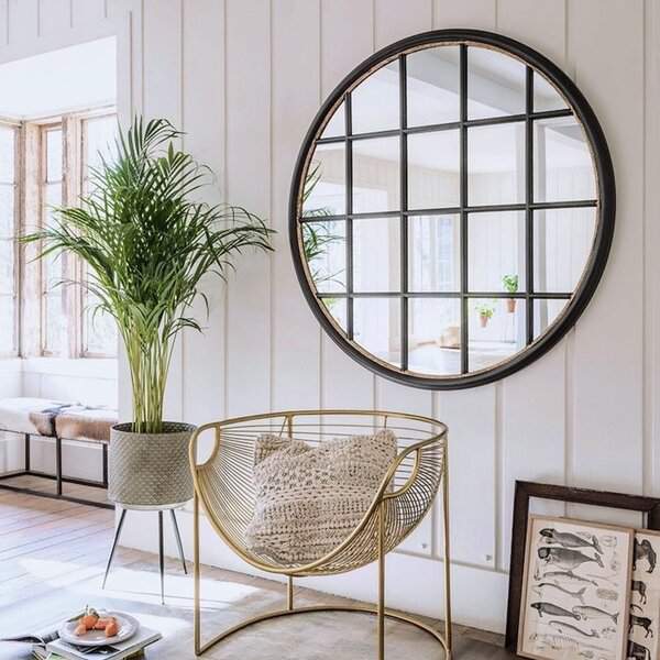 Round grid-framed mirror placed opposite a window, reflecting light in a bright living space with gold accents, greenery, and textured decor.