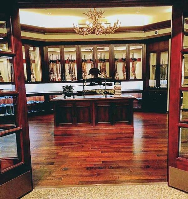 A grand gun room featuring floor-to-ceiling wooden cabinetry, a central display island, and an antler chandelier for a luxurious collector’s ambiance.