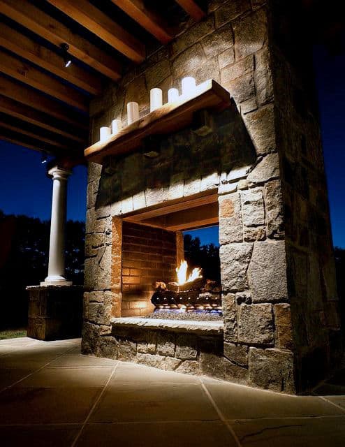 Outdoor stone fireplace with a lit fire and candles on the mantel, under a wooden pergola at night