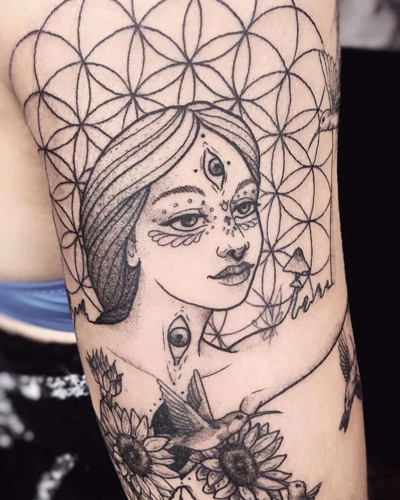 Tattoo of a mystical woman with a third eye, surrounded by geometric patterns, sunflowers, hummingbirds, and fine dotwork details.