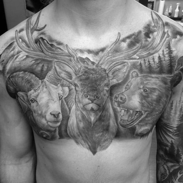 Realistic black and grey wildlife chest tattoo featuring an elk, a ram, and a bear, with detailed shading and a mountainous forest background.
