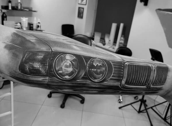 Realistic BMW front grille and headlights tattoo on forearm, featuring detailed shading for a bold and lifelike automotive design.