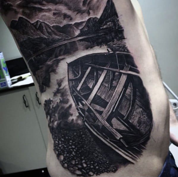 Realistic side tattoo of an overturned wooden boat on a rocky shore, with still lake waters and distant mountains under a moody sky.