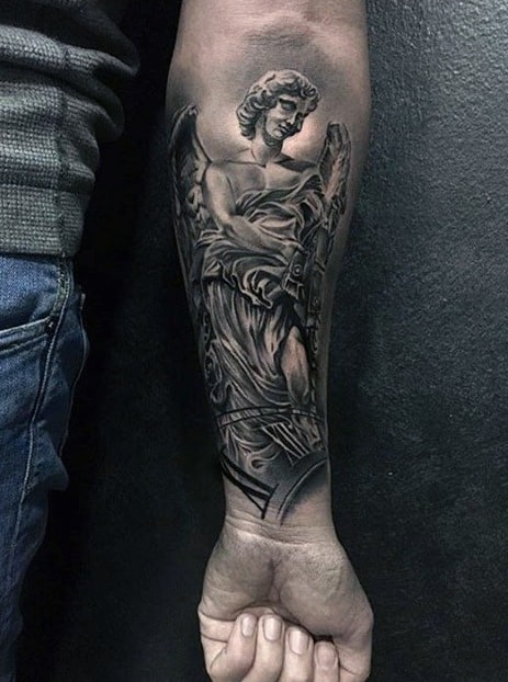 A detailed black and grey tattoo on the forearm featuring an angel holding a sword, with intricate shading and a dramatic pose.