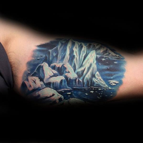 Realistic iceberg tattoo on the upper arm with glowing blue tones, rocky foreground, and a starry night sky over calm waters.