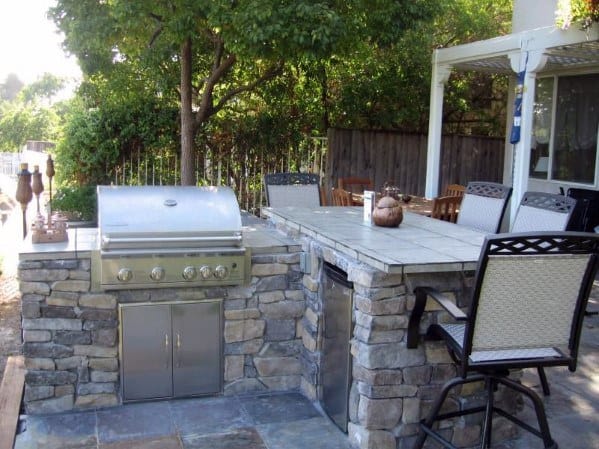 Outdoor kitchen with built-in grill, stone countertop, and patio seating under a leafy tree