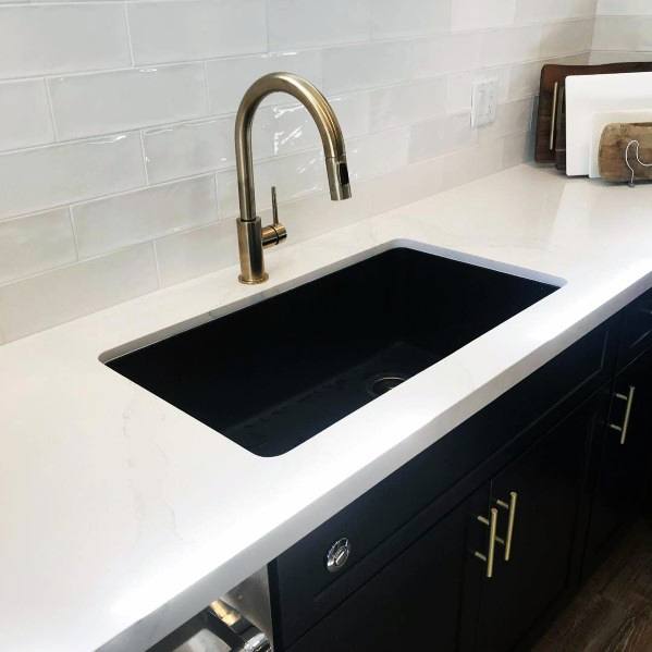 Black kitchen sink with gold faucet, white countertop, and black cabinets with gold handles.