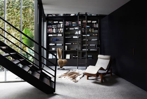 Modern room with a staircase, large bookshelf, chair, and rug; bright light from tall windows, lush plants outside