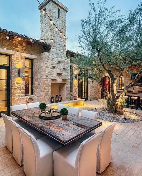 Charming patio with brick accents, a wooden table, white chairs, and an olive tree