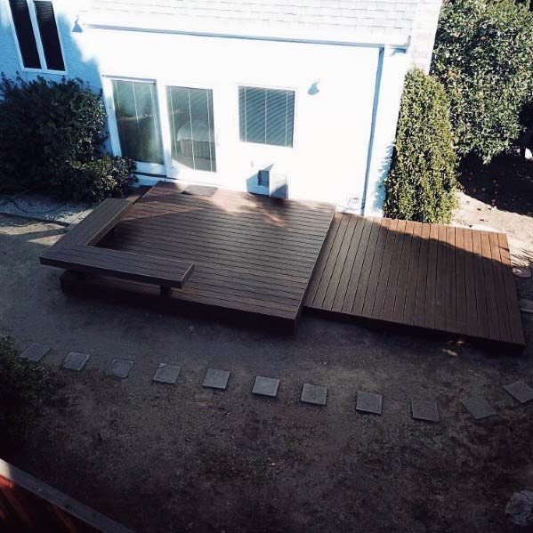 Wooden deck with built-in bench in a lush backyard, adjoining a white house