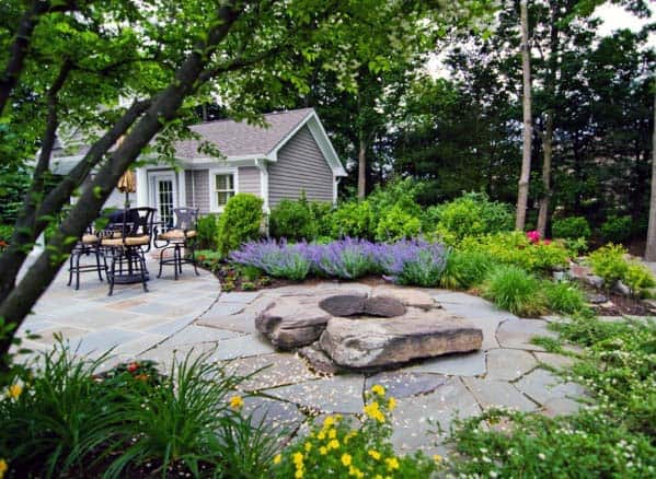 A lush garden with a stone patio, outdoor dining set, and a small house surrounded by trees and vibrant flowers