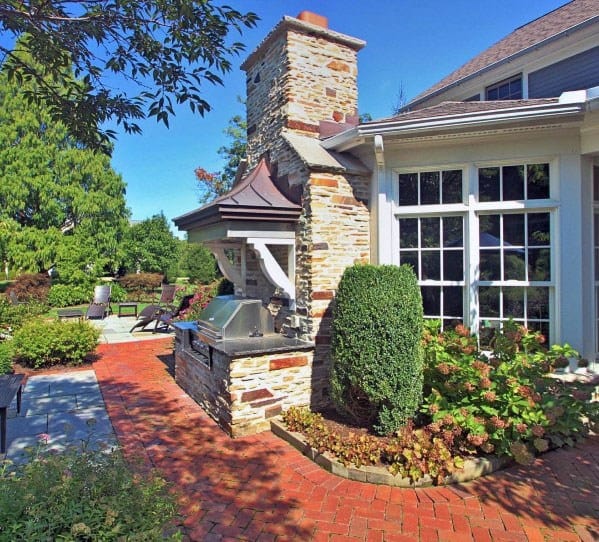 Brick walkway leading to a stone outdoor kitchen with a grill and chimney, surrounded by lush greenery and patio seating