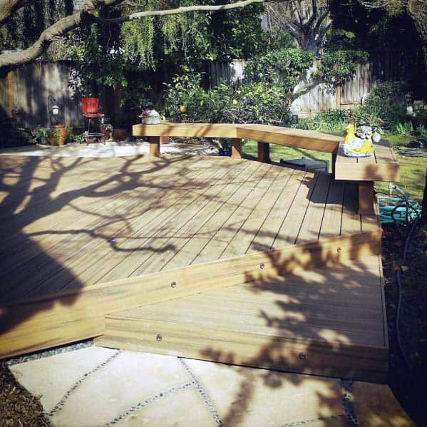 Curved wooden bench on a deck, nestled among trees and garden plants in a serene backyard setting