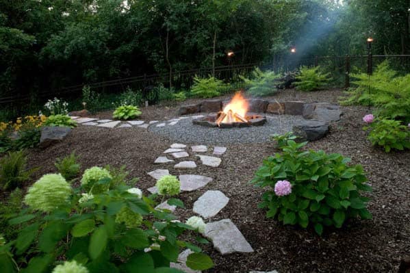 A garden with a stone path leads to a lit fire pit surrounded by greenery and flowers, set against trees