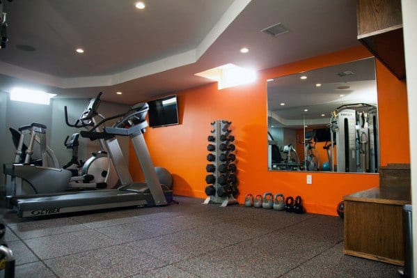 Home gym with orange accent wall, rubber flooring, treadmill, elliptical, and weight rack.