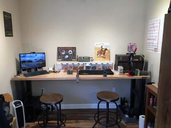 Modern reloading bench with a clean wooden surface, dual stools, mounted press, and a computer for added functionality