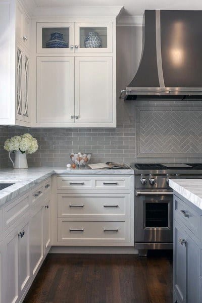 Modern kitchen with white cabinets, gray herringbone backsplash, and stainless steel range hood.