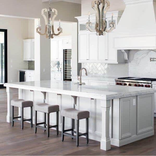 White kitchen with marble island, upholstered bar stools, and elegant chandeliers.