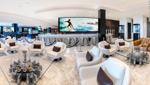 Luxurious living room with white chairs, glass tables, and a large screen displaying a surfer in action